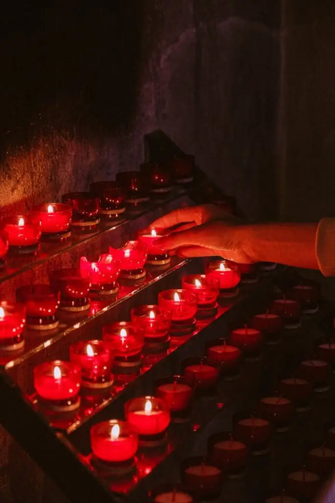 A serene scene of red votive candles being lit by a hand inside a dimly lit church.